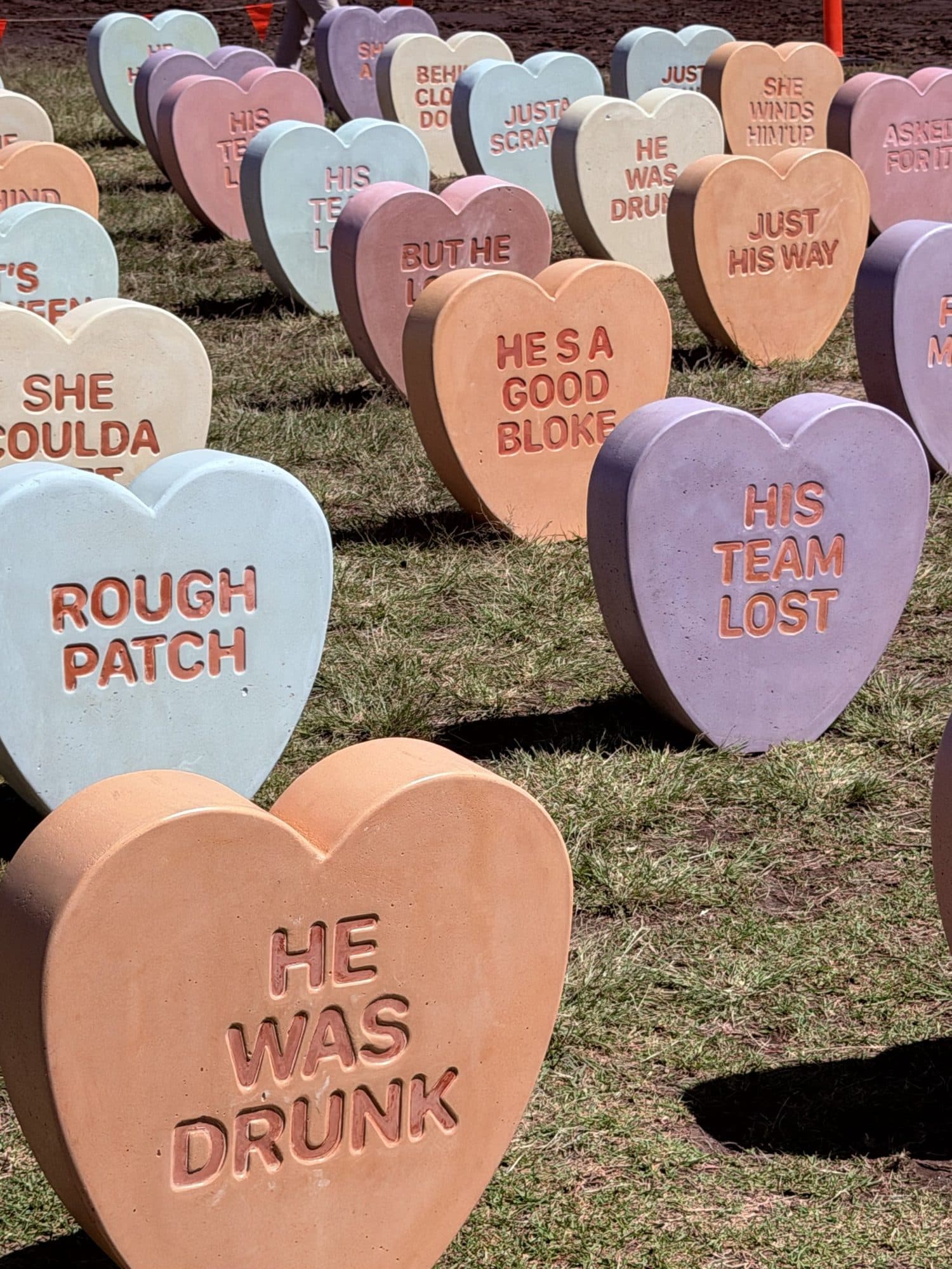 Rows of pastel concrete hearts in the grass at Marks Park, carved with phrases including He was drunk, Rough patch, He's a good bloke and His team lost