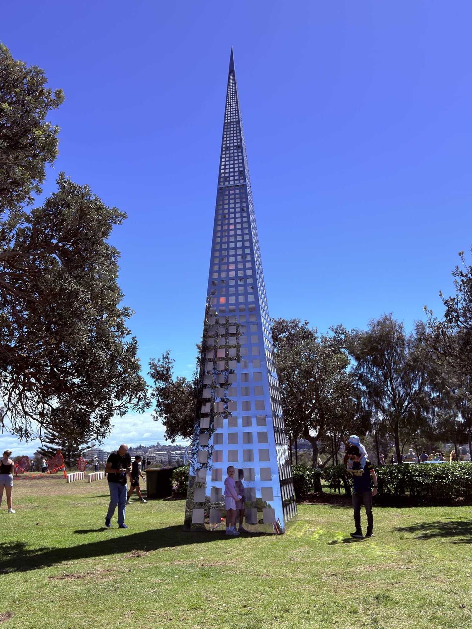 Clayton Blake's Pinnacle: a tall, narrow, mirrored steel spire with a grid of square cut-outs, visitors standing at its base on the Marks Park lawn