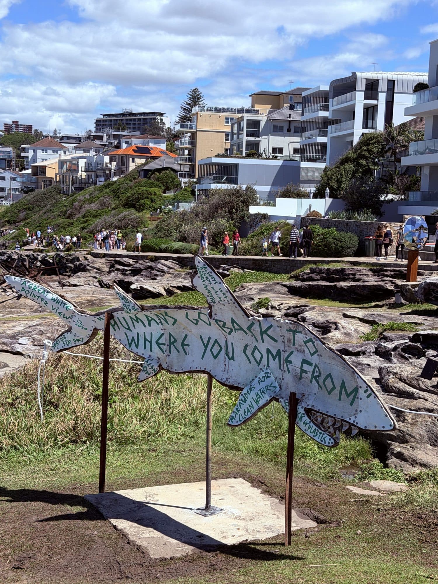 Ian Swift's driftwood great white shark on poles, with hand-painted lettering along its body, above the rocks with Bondi apartments behind